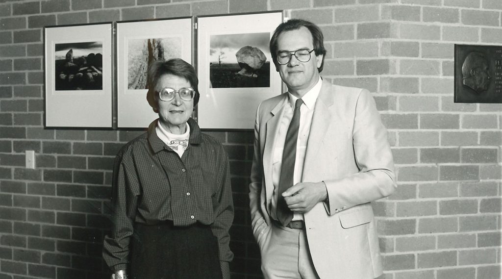 Black and white image of a man and a woman standing in front a brick wall. 