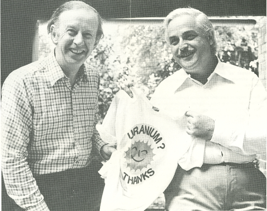 Black and white image of two men smiling. One is holding a tshirt that reads, 'Uranium? No thanks'. 