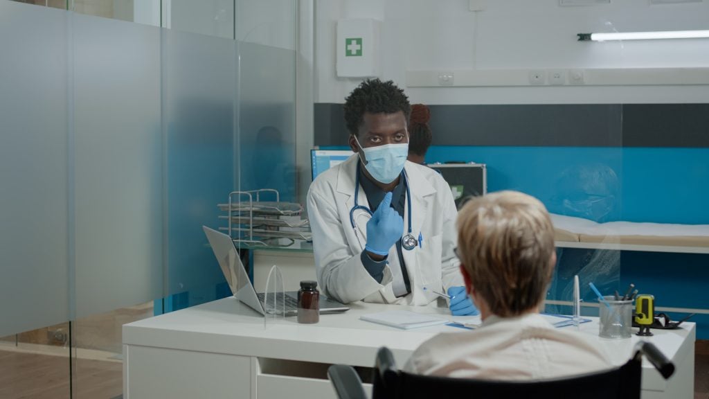 A doctor in a mask and gloves sits at a desk in a hospital, talking to a patient in a wheelchair.