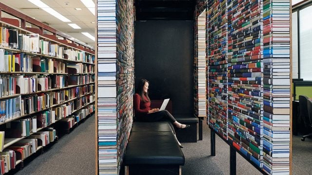 Student sits on a bench surrounded by books, typing on her laptop