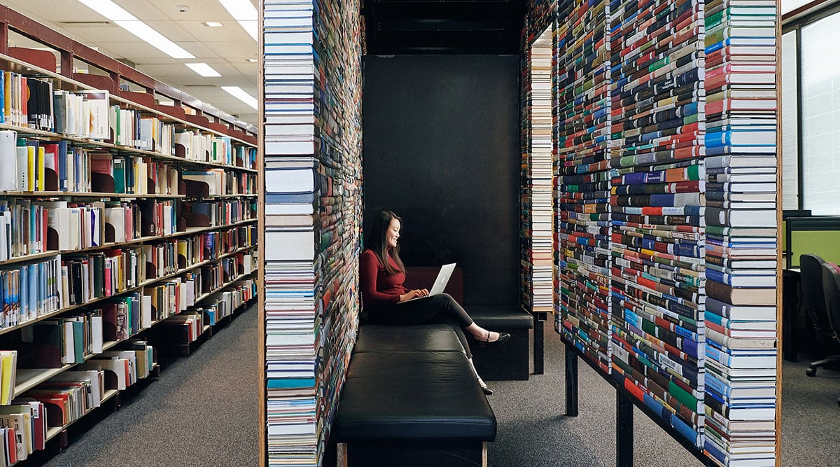 Student sits on a bench surrounded by books, typing on her laptop