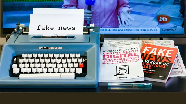 A typewriter and a stack of books on a desk, with a television in the background. The typewriter paper and book covers say 'fake news'.