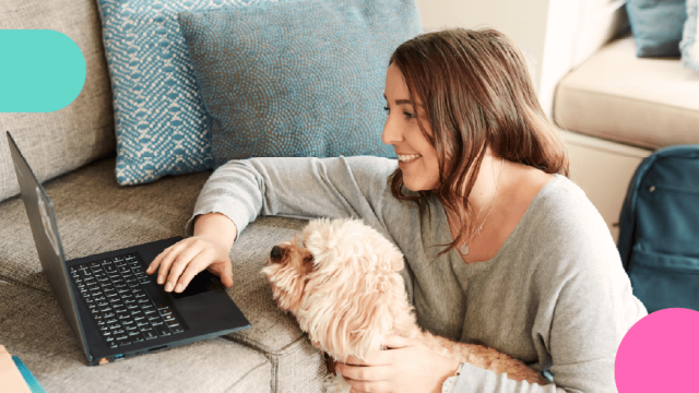 Woman sitting next to her couch, looking at her laptop with her dog beside her.