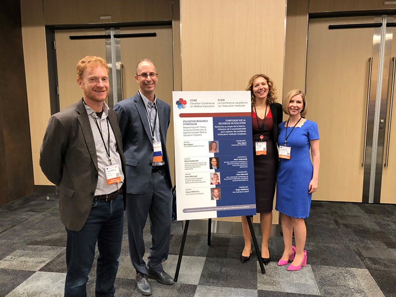 Photograph of A/Prof. Angus McMurtry, A/Prof. Mark Goldszmidt, Dr Rola Ajjawi and A/Prof. Anna McLeod with a promotional board for their symposium.