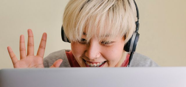 A woman wearing headphones smiling and waving at laptop screen