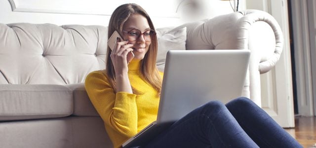 woman smiling as she talks on phone while sitting on floor with laptop in living room