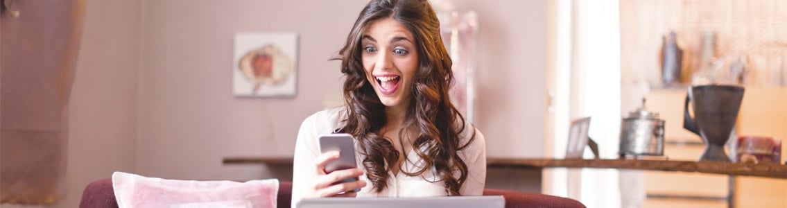 Woman looking surprised and happy while looking at phone and holding laptop