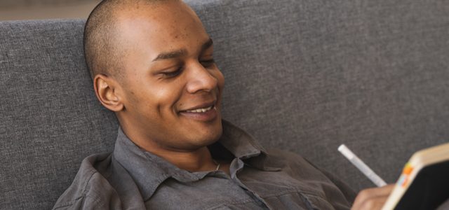 man smiling as he sits on couch writing in notebook
