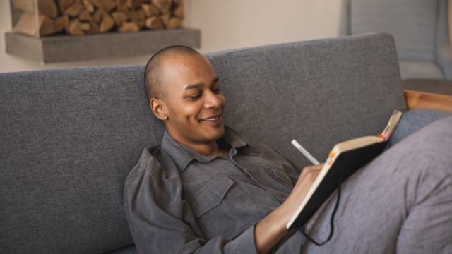Man smiling as he writes in notebook at home on couch