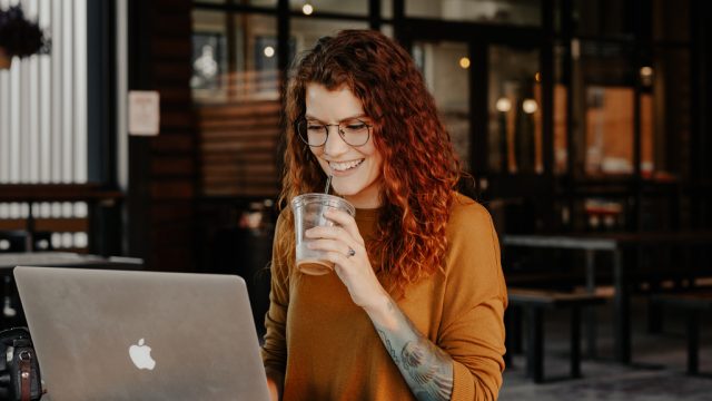 Woman smiling as she sips drink and works on laptop in cafe