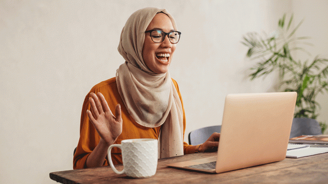 Smiling young woman in hijab waving at laptop screen