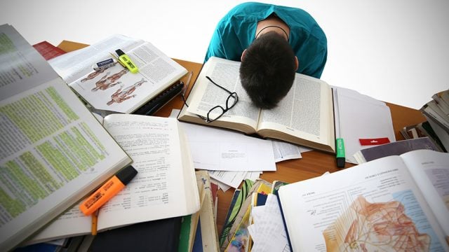 Student resting head on pile of notes and books