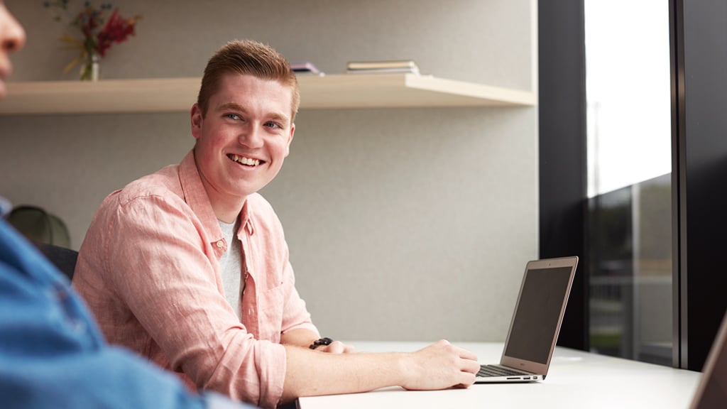 Student smiles at friend as he works on laptop in study space