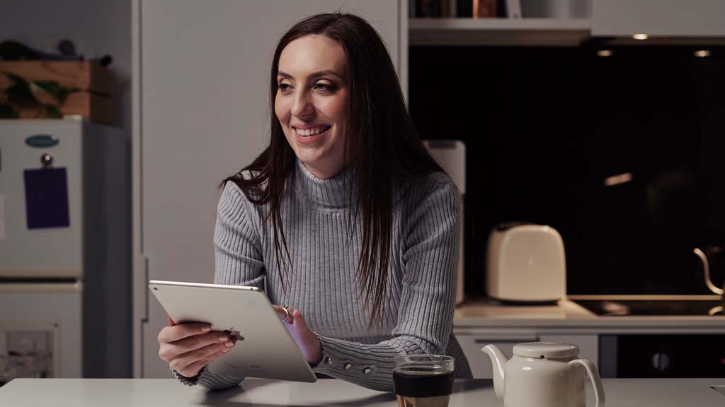 Student smiling as she holds tablet while seated at kitchen bench at home