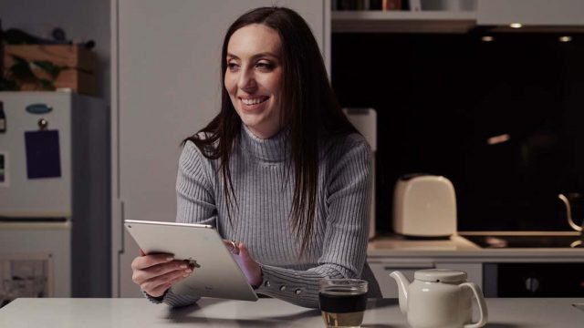 Student smiling as she holds tablet while seated at kitchen bench at home