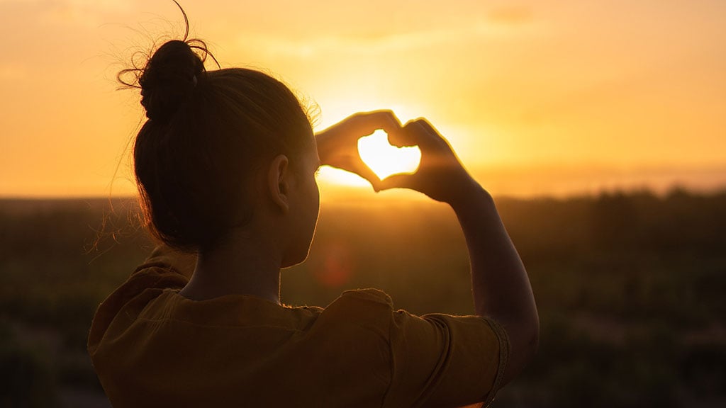 Women at sunset with hands in heart shape framing sun