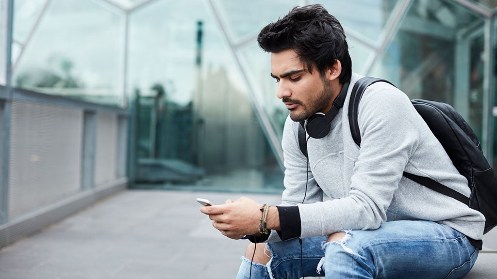 Male student wearing backpack and headphones and looking at phone