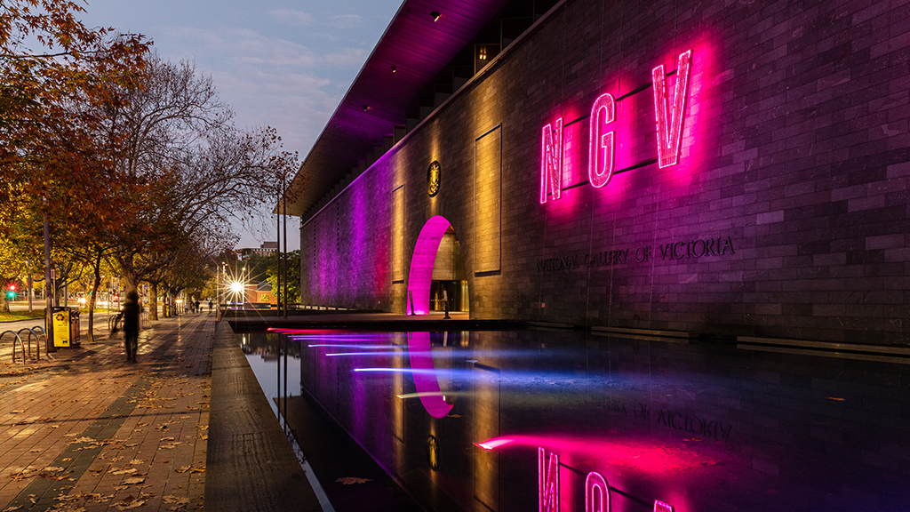 The sign and fountain outside the NGV lit in neon in the evening