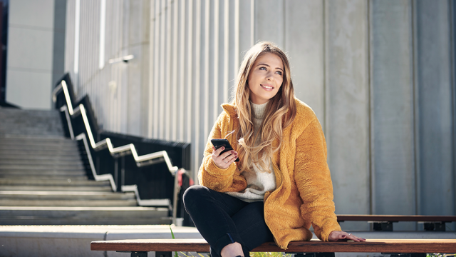 A student sitting on the stairs on campus holding a phone and smiling that they still have wi-fi access