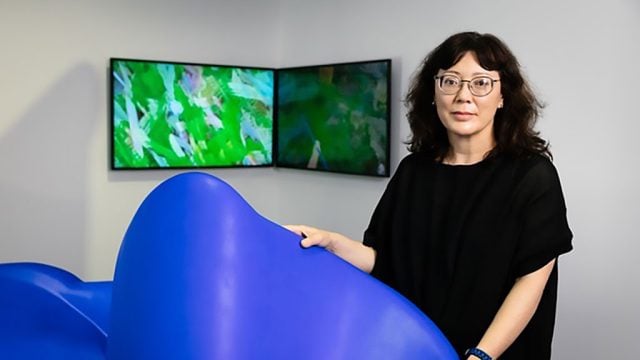 Artist Fayen d’Evie: ID: A woman with black wavey hair and wearing eyeglasses and a black tunic style dress looks at the camera smiling pensively. She is holding a section of a wavey plastic blue sculpture in her hands. Behind her are two flat screens with blurry green images on them. 