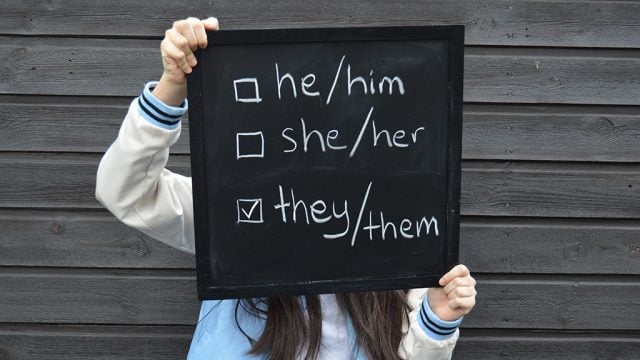 Person holding chalkboard with pronoun options with checkboxes. 'They/them' is ticked.