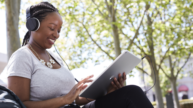 A woman wearing headphones sits on a bench, focused on her tablet, enjoying some quiet time outdoors.