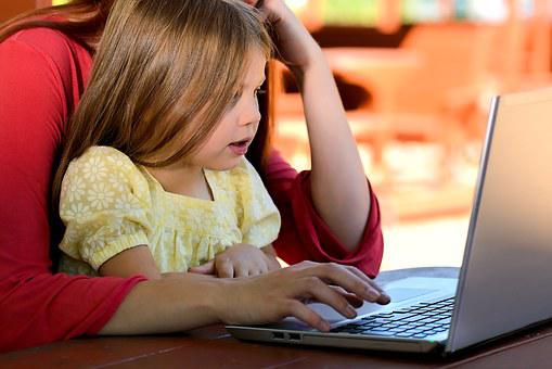 An adult operates a laptop while a young girl sits on their lap, looking at the screen.
