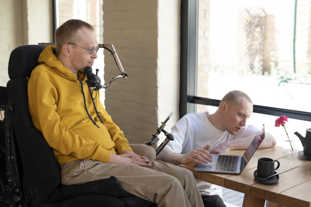 Two men work together on a laptop. One man is crouching down and the other is seated in a supportive wheelchair and uses a joystick with his chin.