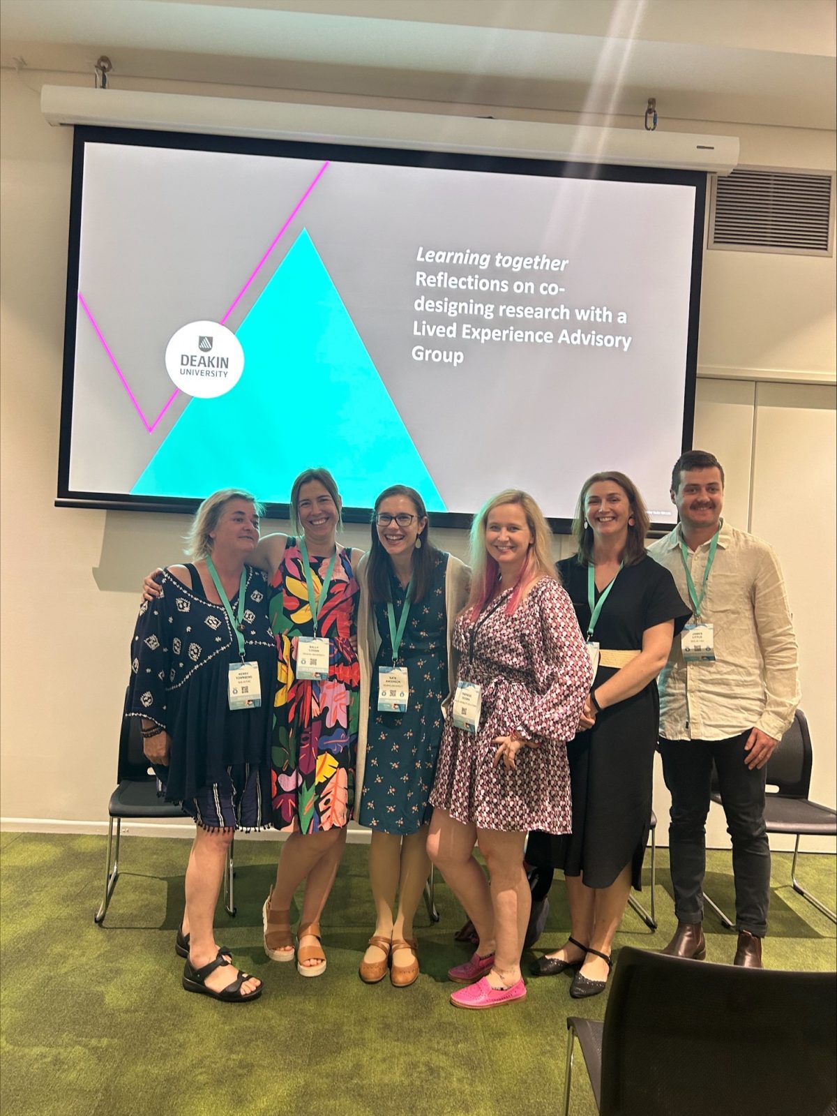 Team members Kerry Townsend, Sally Logan, Kate Anderson, Therese Dogra, Valerie Watchorn, and James Little are smiling and posing in front of a slide titled "Learning Together: Reflections on co-designing research with a Lived Experience Advisory Group", displayed on a large digital presentation screen.