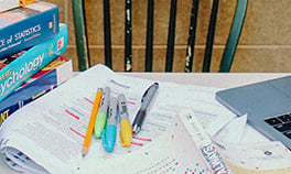Photograph of assorted pens and pencils on marked-up papers with book in foreground